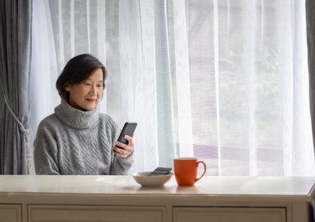 Senior woman smiling and reading smartphone. A coffee cup and dark chocolate on white table.の写真素材