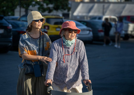 Senior woman walking using a mobility walker across the carpark, accompanied by her daughter on a...