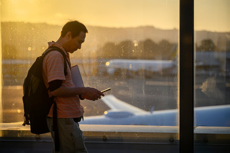 Man carrying a laptop and checking his mobile phone at the airport. Out-of-focus airplanes parked outside the terminal window at sunset.の写真素材