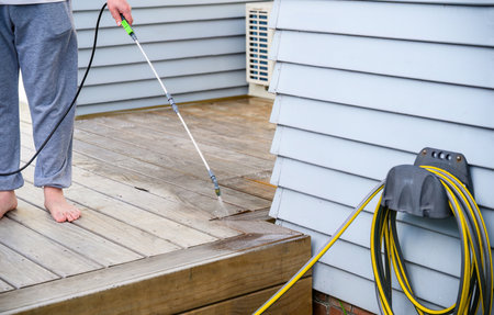 Man cleaning wooden deck with hand-held pressure sprayer. Garden hose in the foreground.の写真素材