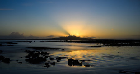 Sunbeams glowing at the crest of Rangitoto Island as the sun is rising. Milford Beach. Auckland.の写真素材