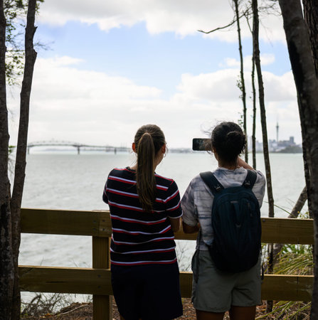 Woman taking photos of Auckland Harbour Bridge and Sky Tower using smartphone. Kauri Point Centennial loop track. Vertical format.の写真素材
