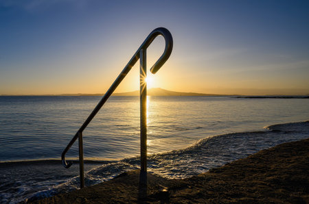Handrail framing sunrise over the top of Rangitoto Island. Milford Beach. Auckland.の写真素材