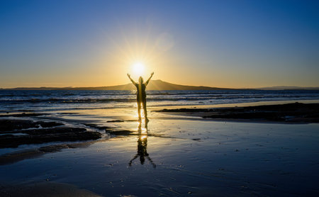 Woman raises arms towards the rising sun. Sunrise over the Rangitoto Island. Milford Beach. Auckland.の写真素材