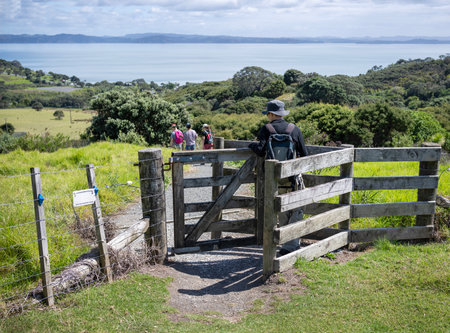 People walking through the farm gate on a hiking track. Electric  Fence sign hanging on the fence. Shakespear Regional Park. Auckland.の写真素材
