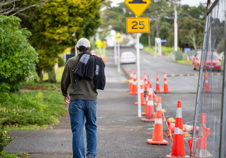 Man walking on a pedestrian footpath. Orange traffic cones and metal fences on the road. Road works...