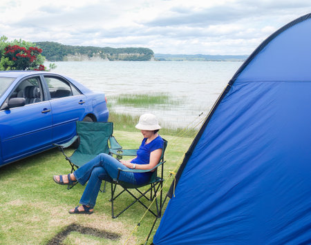Woman sitting on the camping chair and scrolling on her smartphone by the tent. Camping in summer. Waikato. New Zealand.の写真素材