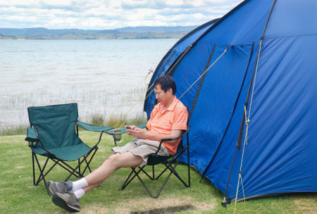 Man sitting on the camping chair and scrolling on his smartphone by the tent. Camping in summer. Waikato.の写真素材