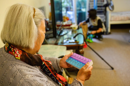 Senior woman holding a pill box inside home.  Out-of-focus adult daughter cleaning the house.の写真素材