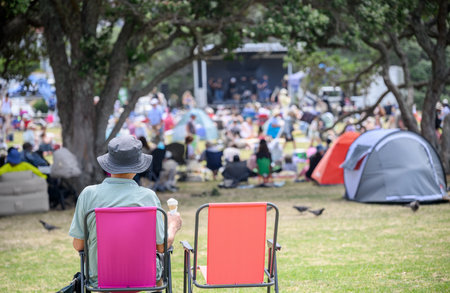Man sitting on camping chair with an empty chair beside. Unrecognizable crowds listen to the music concert in the park.の写真素材