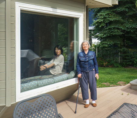 Elderly woman posing for photos, adult daughter smiling from inside the bay window. Happy family holiday.の写真素材