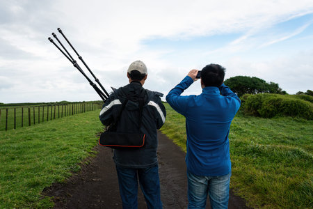 Two men walking on a rural road. One man taking smartphone photos. Another man holding tripod....