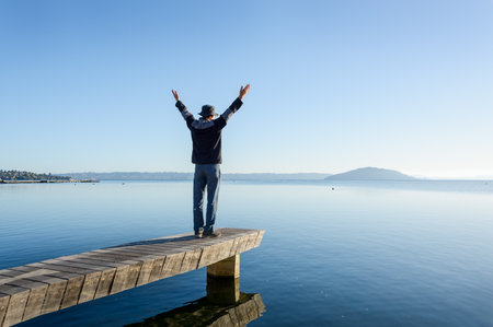 Man standing on the jetty, arms raised towards Mokoia Island. Lake Rotorua. New Zealand.の写真素材