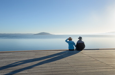 Couple sitting on the lakefront boardwalk by Lake Rotorua. Mokoia Island in the distance. Rotorua.の写真素材