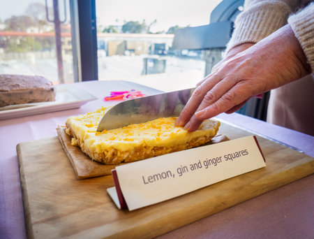 Hands of a woman cutting a cake. Cardboard with words âLemon, gin and ginger squaresâ.の写真素材