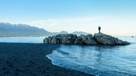 Woman standing on the rocks at the beach. Snow-capped mountains in the distance. Kaikoura. New Zealand.の写真素材