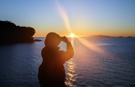 Woman taking photos at sunrise. Sun starburst shing above the horizon. Auckland.の写真素材
