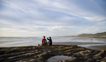 Couple sitting on rocks and watching sunset at Muriwai Beach. Auckland.の写真素材