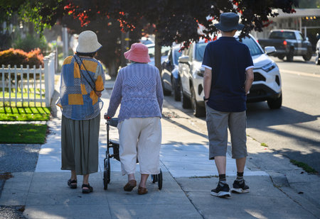 Rear view of an elderly woman walking using a mobility walker on the pedestrian footpath, accompanied by her adult children. California.の写真素材