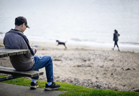Man reading his phone on the picnic bench by the beach. Unrecognizable people walking the dog on the...