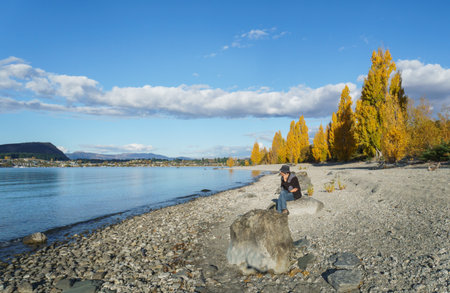 Tourist sitting on the rock and making a phone call. Golden autumn trees along Lake Wanaka. South Island. New Zealand.の写真素材