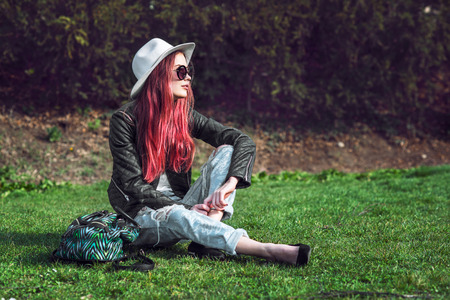 Beautiful stylish red haired fashion hipster model woman sitting outdoors on green grass at park wearing sunglasses, hat and black leather coat.の写真素材