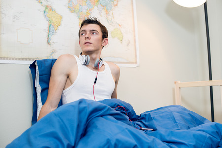 Young man woke up and listening music wearing headphones in the bed at home. Handsome student man sitting in the bed wearing white t-shirt.の写真素材