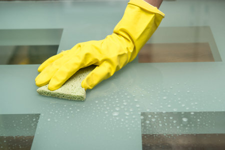 Close-up photo of female hand washing glass table in with cleaning spray and sponge wearing gloves...