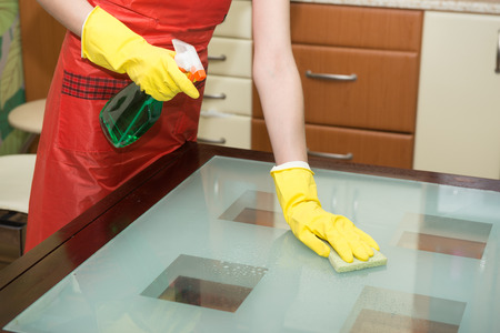 Housekeeper washing glass table in with cleaning spray and sponge wearing gloves in apartment.の写真素材