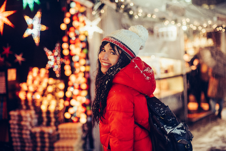 Beautiful young smiling woman enjoy snow winter time on Christmas fair in night city wearing hat and red jacketの写真素材