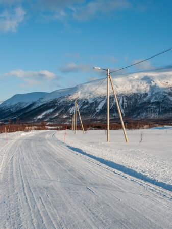 Snowy road with lamp posts and mountains in the background in northern Norway in winterの写真素材