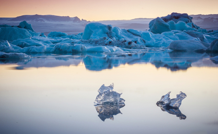 Two small pieces of ice flowing and reflecting in the cold lake with a big iceberg behind, jokulsarlon glacier lagoon, Icelandの写真素材