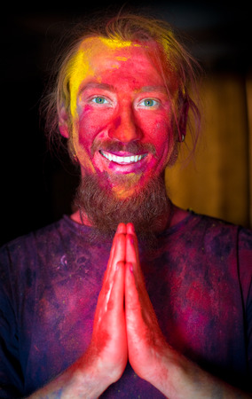 Bright and colorful portrait of the man in paint after Holi celebration. His hands are folded together in "namaste" gesture.の写真素材
