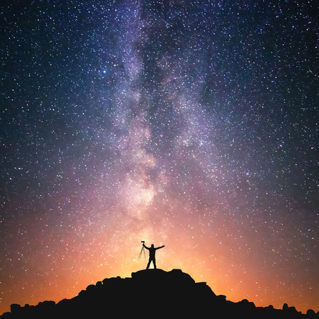 The Man and the Universe. A person is standing on the top of the hill next to the Milky Way galaxy with a tripod in his hands.