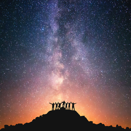 Together we stand. A group of people are standing on the top of the hill next to the Milky Way galaxy holding hands.の写真素材