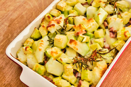 Gratin pattypan squash and zucchini with cheese, ham and rosemary herb in red bowl preparingの写真素材