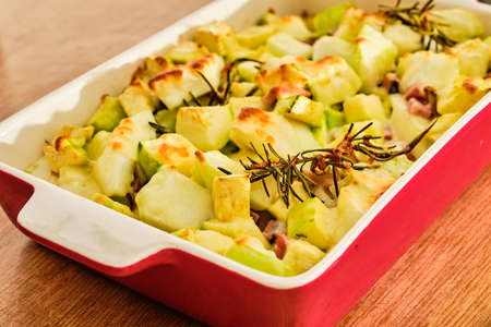 Gratin pattypan squash and zucchini with cheese, ham and rosemary herb in red bowl preparingの写真素材