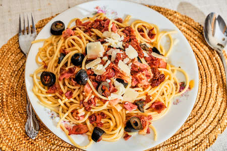 Tasty spaghetti on plate with dried ham, dried tomatoes, olives and parmesan cheese on white plate and fork with spoon servingの写真素材
