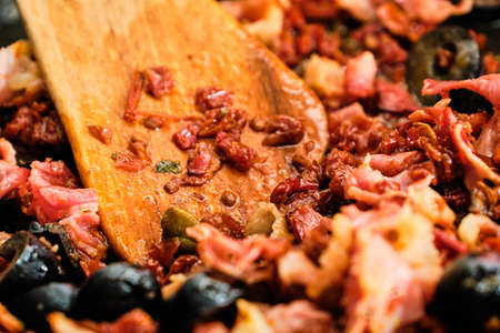 Cooking tasty mixture for spaghetti - dried tomatoes, black olives and dried ham on frying pan preparing with wooden spatulaの写真素材