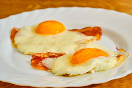 Two tasty fried egg with bacon on wihte plate on wooden tableの写真素材