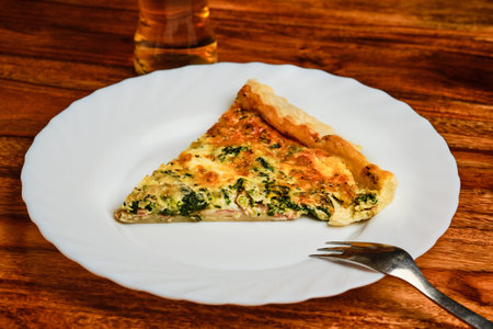 Tasty spinach pie with cheese and ham on white plate with fork on wooden table with beerの写真素材