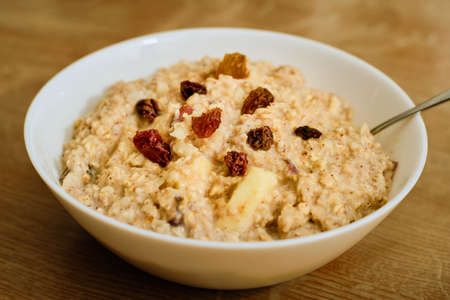 Tasty oatmeal porridge with apples and raisins in white bowl on wooden tableの写真素材