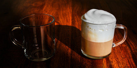 Hot coffee with white foam in glass cup and empty glass on wooden tableの写真素材