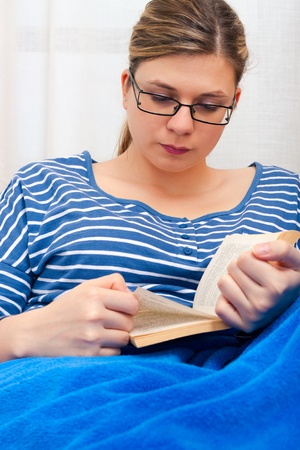Young woman reading book and lying on sofa with blue blanket.の写真素材