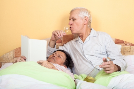 Funny senior couple relaxing in bed with alcohol and bookの写真素材