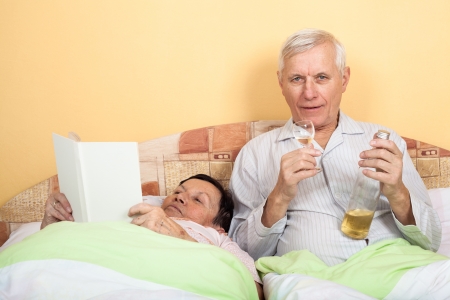 Senior couple resting in bed with alcohol and bookの写真素材