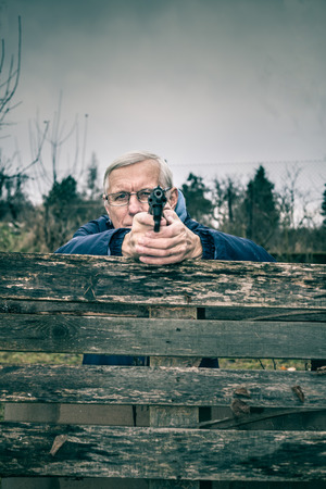 Senior man aiming a gun at you and hiding behind barricade outdoors.の写真素材