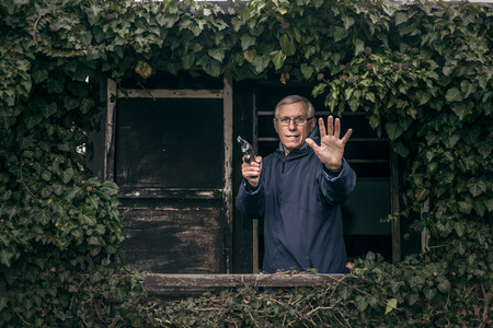 Furious senior man gesturing with a gun protecting his property, standing in front of old overgrown cabin.の写真素材