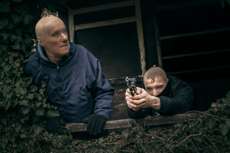 Two masked armed men hiding on overgrown porch of old cabin.の写真素材