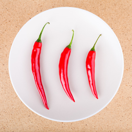 Three fresh raw red hot chili peppers on plate, over light wooden background.の写真素材